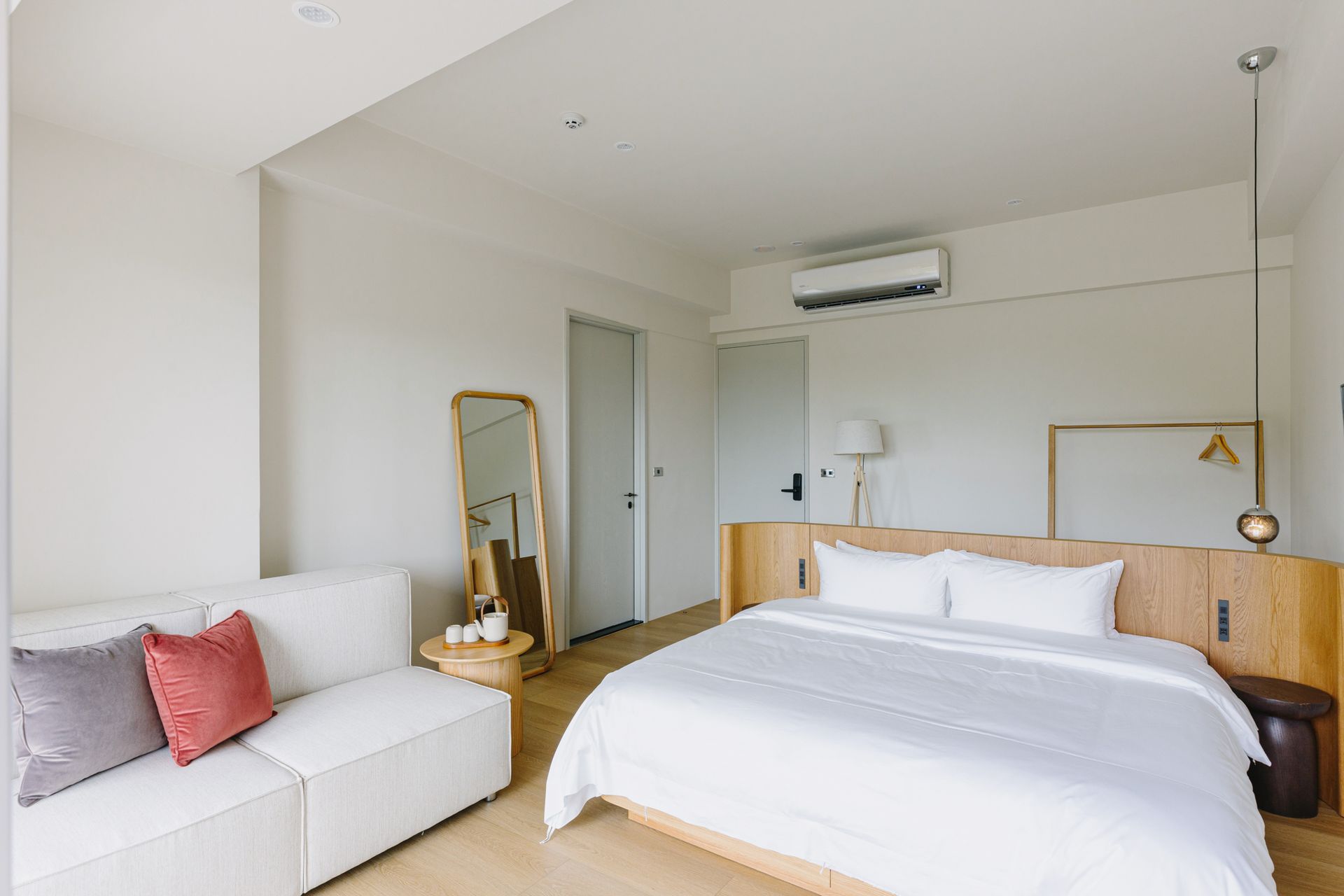 Bright white bedroom with bed, sofa, and full-length mirror. Wooden accents, air conditioner.