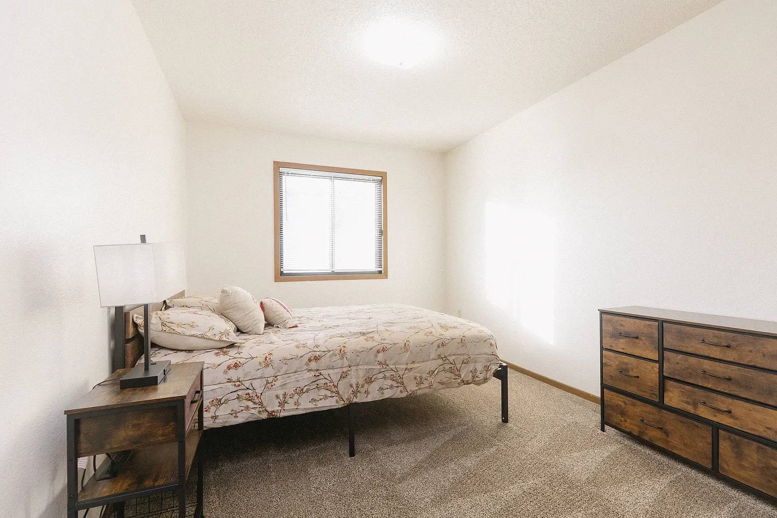 Bedroom with bed, nightstand, and dresser. Neutral tones, window in the back, carpet flooring.