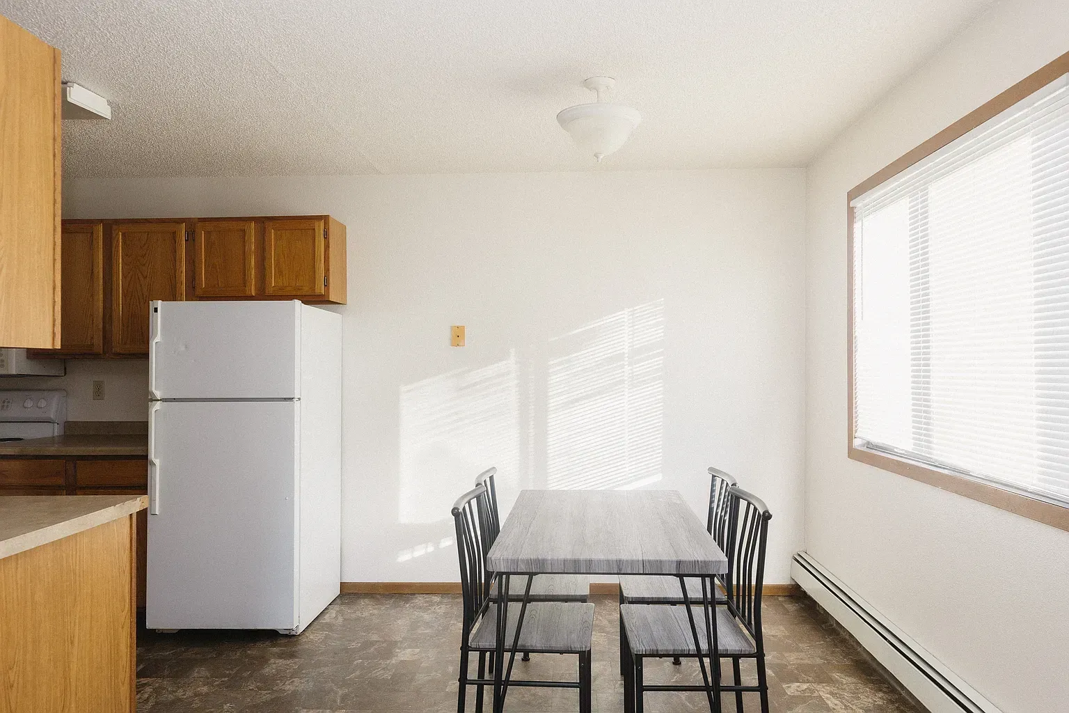 Dining area with table and chairs, refrigerator, and kitchen cabinets. Blinds cover window.