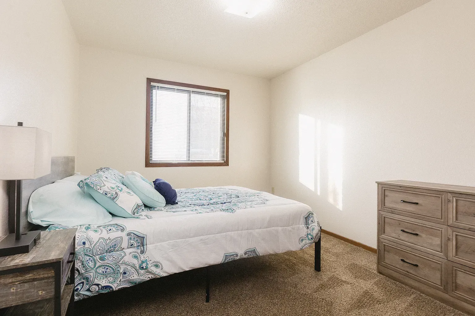 Bedroom with bed, dresser, window, and lamp. Earth-toned carpet and white walls.