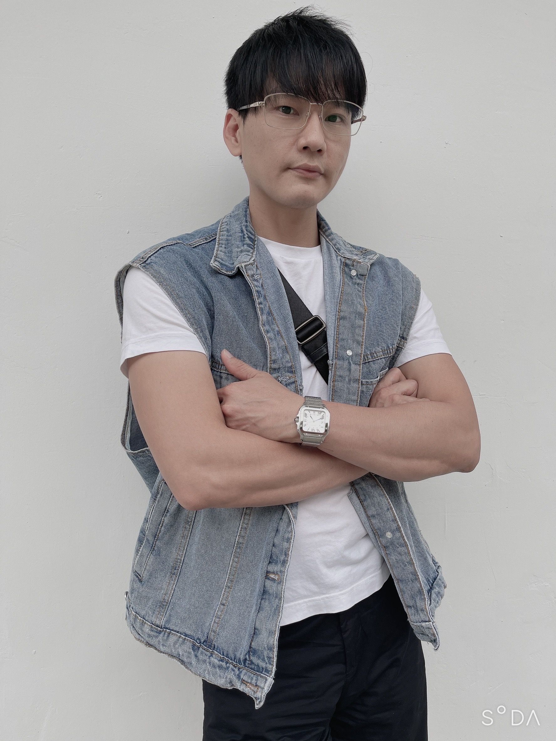 A man wearing a denim vest and glasses is standing with his arms crossed.