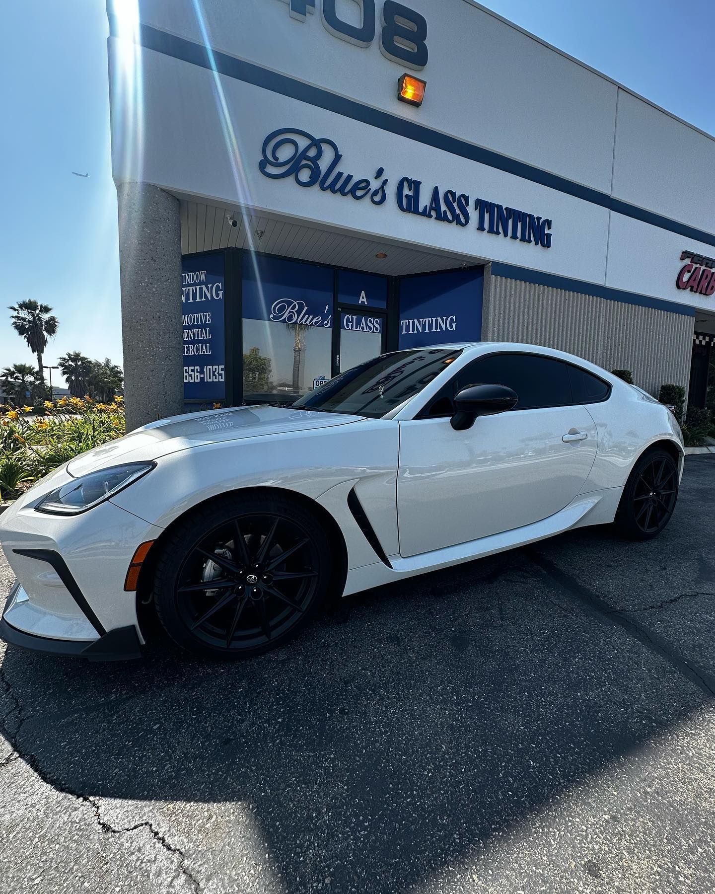 White sports car parked in front of a blue window tinting business on a sunny day.