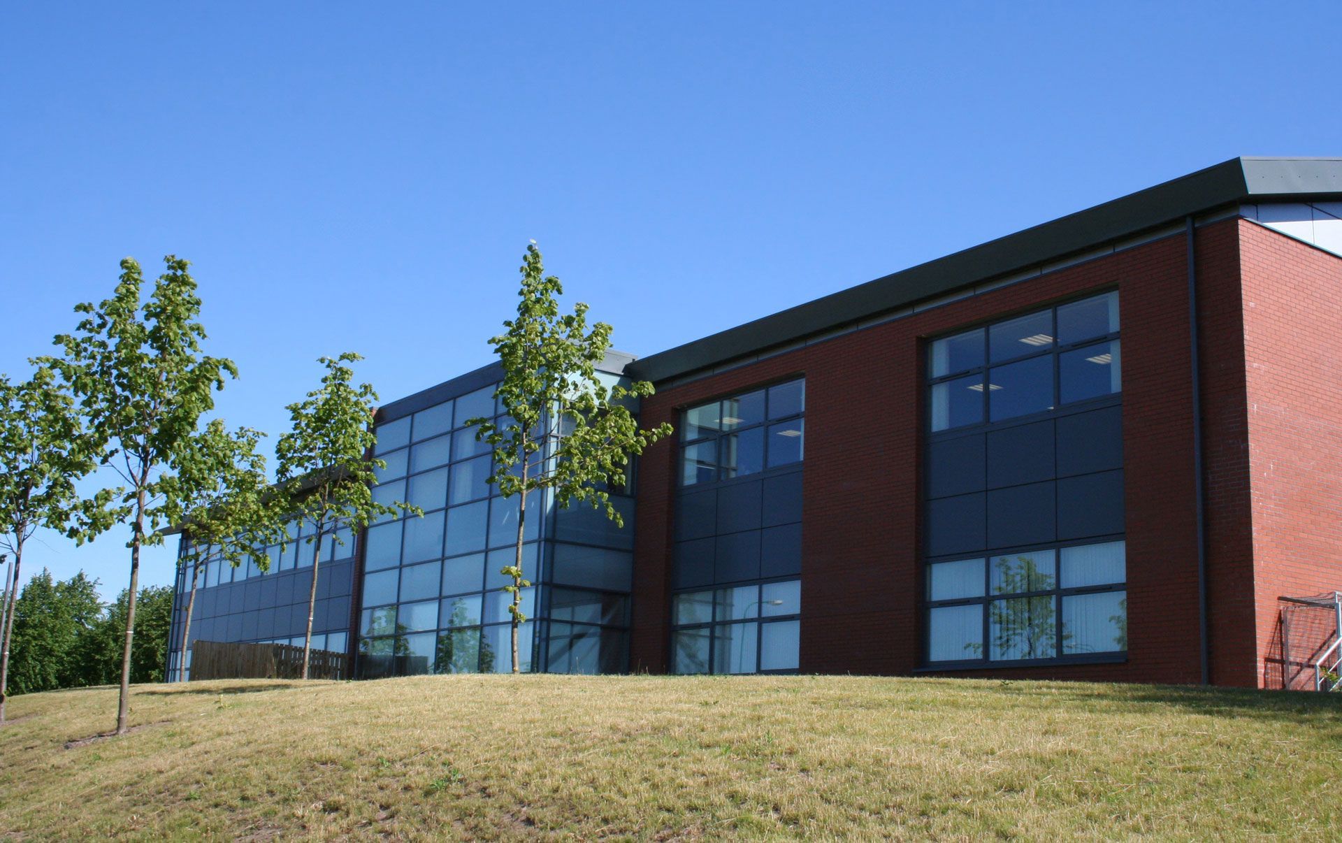Modern brick and glass building with trees on a grassy hill under a blue sky.