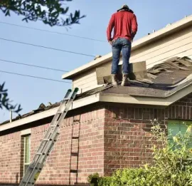 A person wearing a red shirt and blue jeans stands on a sloped residential roof with an extension ladder resting nearby.
