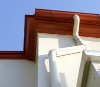 A low-angle view of a building corner with a red roof gutter and a white downspout against a blue sky.