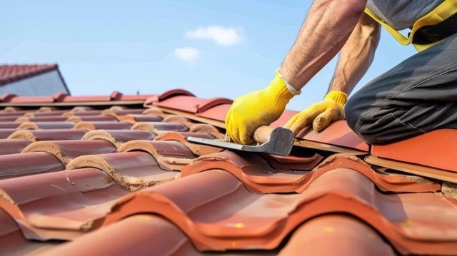 A person wearing yellow work gloves repairs a terracotta tiled roof with a metal tool under a blue sky.