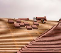 Roof under construction showing rows of wooden battens, stacks of red clay roof tiles, and a power tool on the slope.