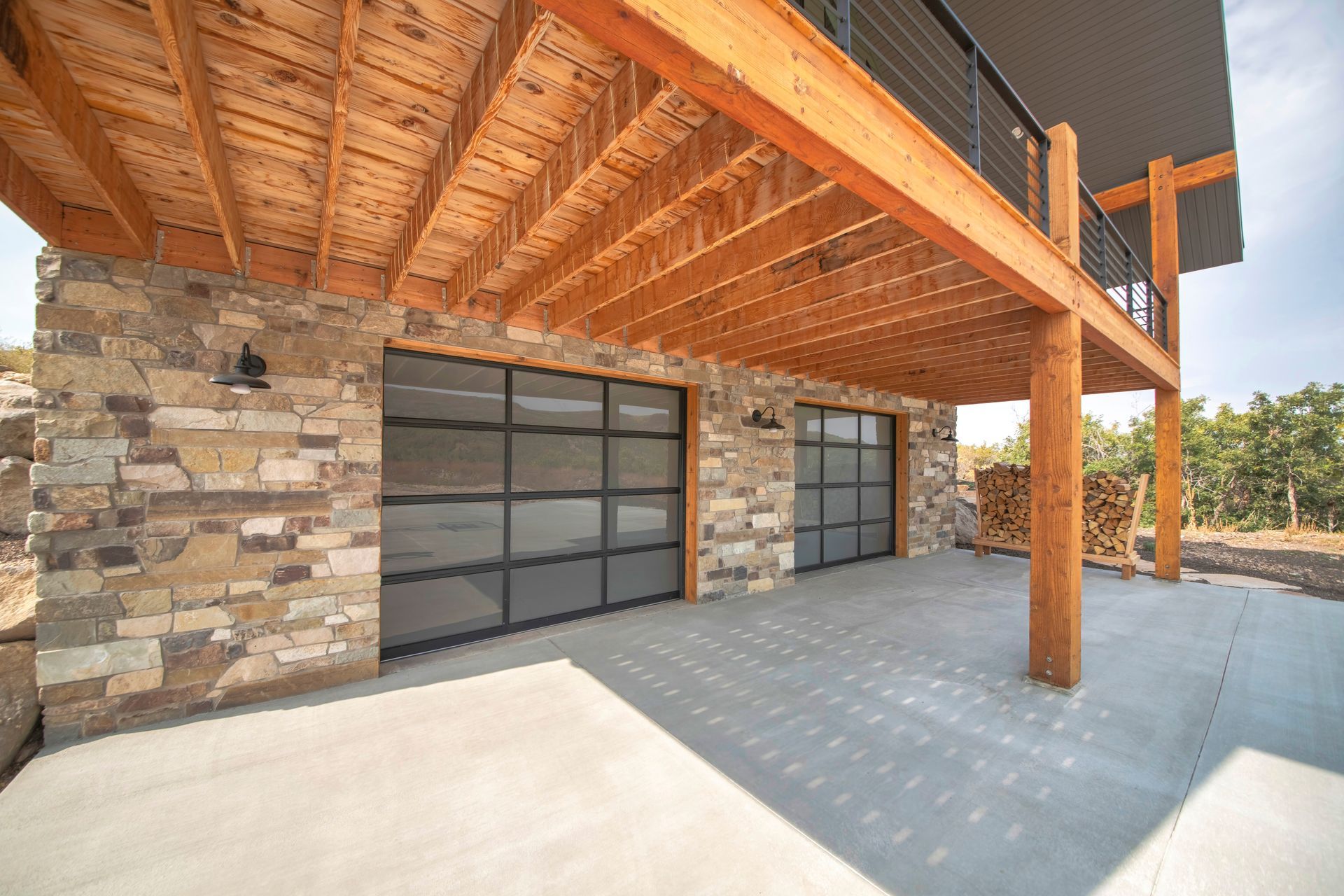 Covered concrete patio under a wooden deck framed by natural stone pillars and glass garage doors