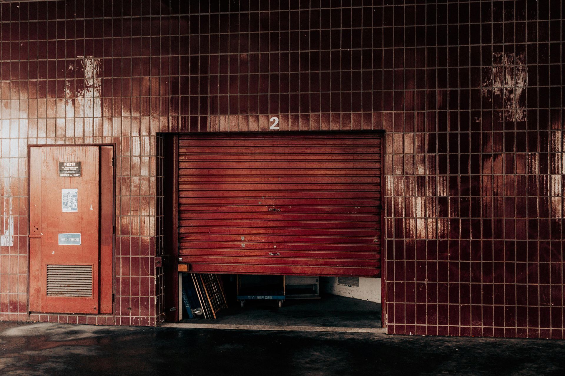 Red-tiled exterior wall with open, red garage door revealing an interior space. A door is on the left.
