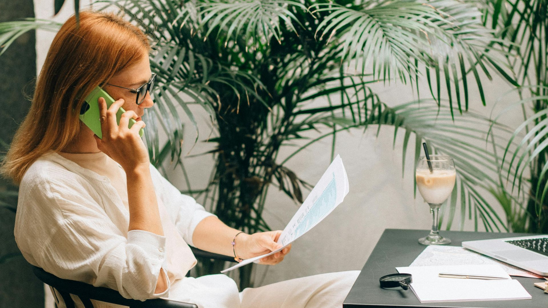 Woman in glasses on a phone call, looking at papers, with a laptop, drink, and plants on a table.