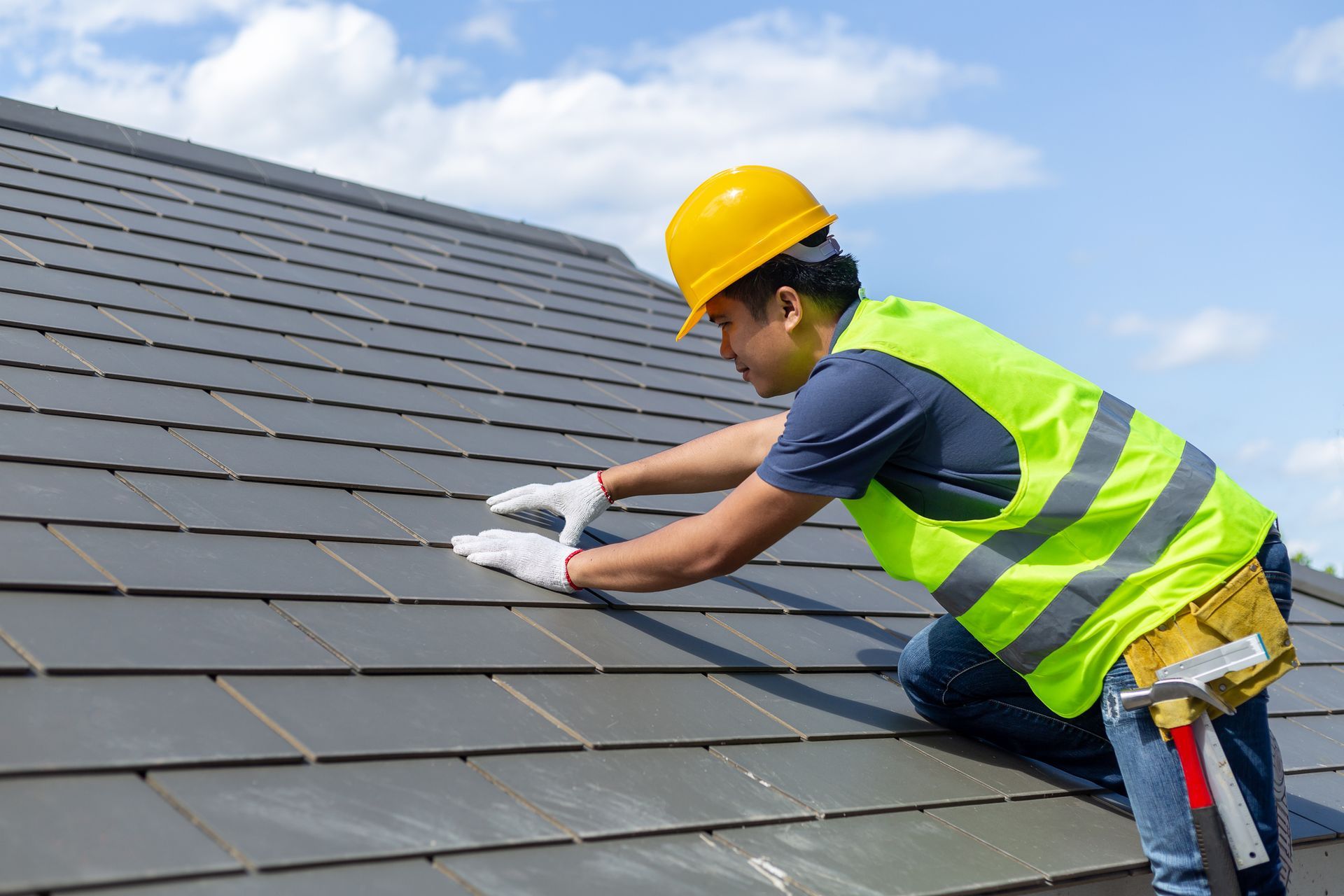 Roofer in yellow hard hat and safety vest, installing dark gray tiles on a roof.