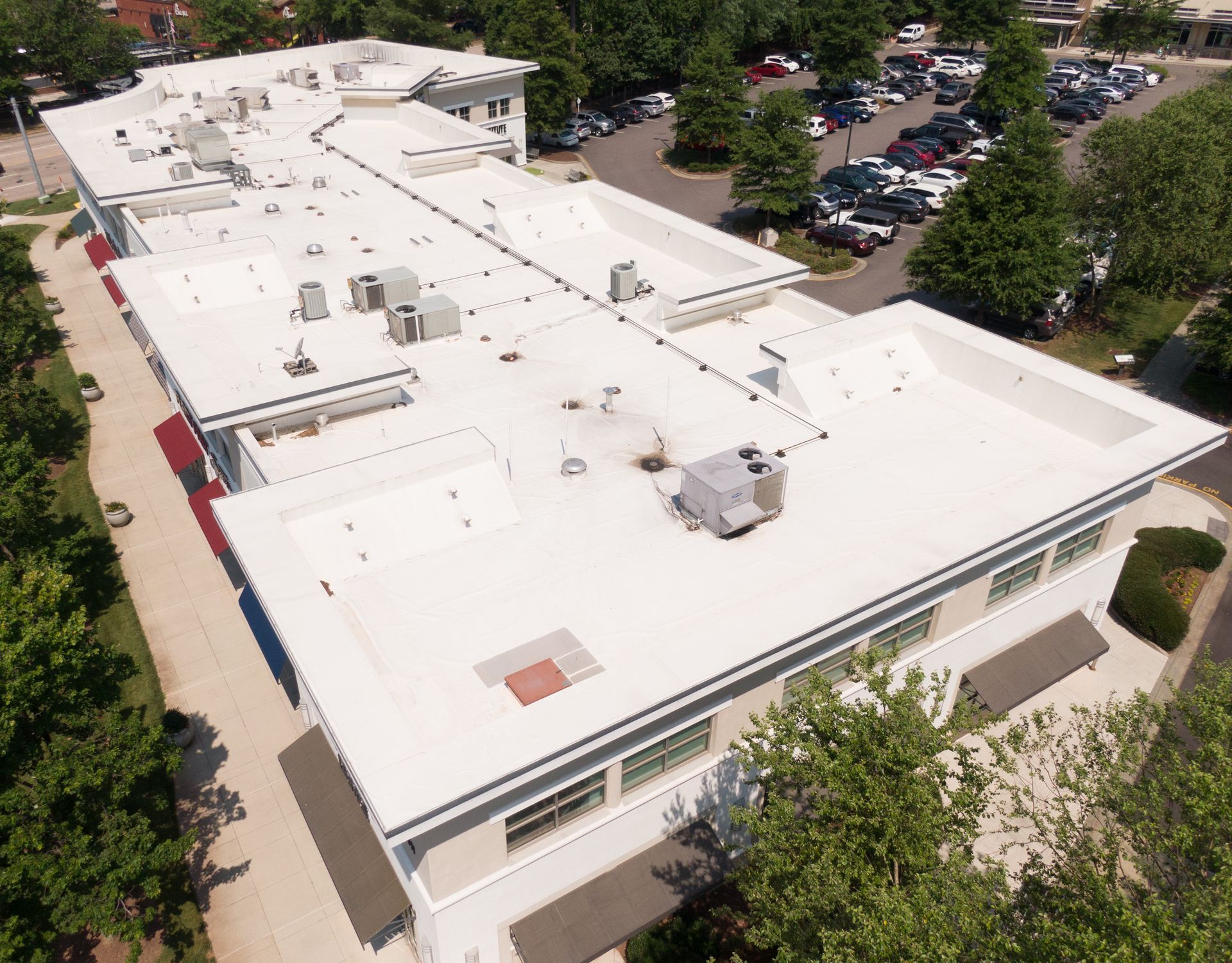 Aerial view of a white commercial building with a flat roof, surrounded by trees and a parking lot.
