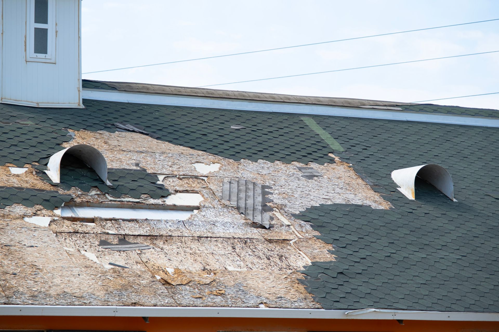 Damaged roof with missing shingles, two metal vents, and a white building section.