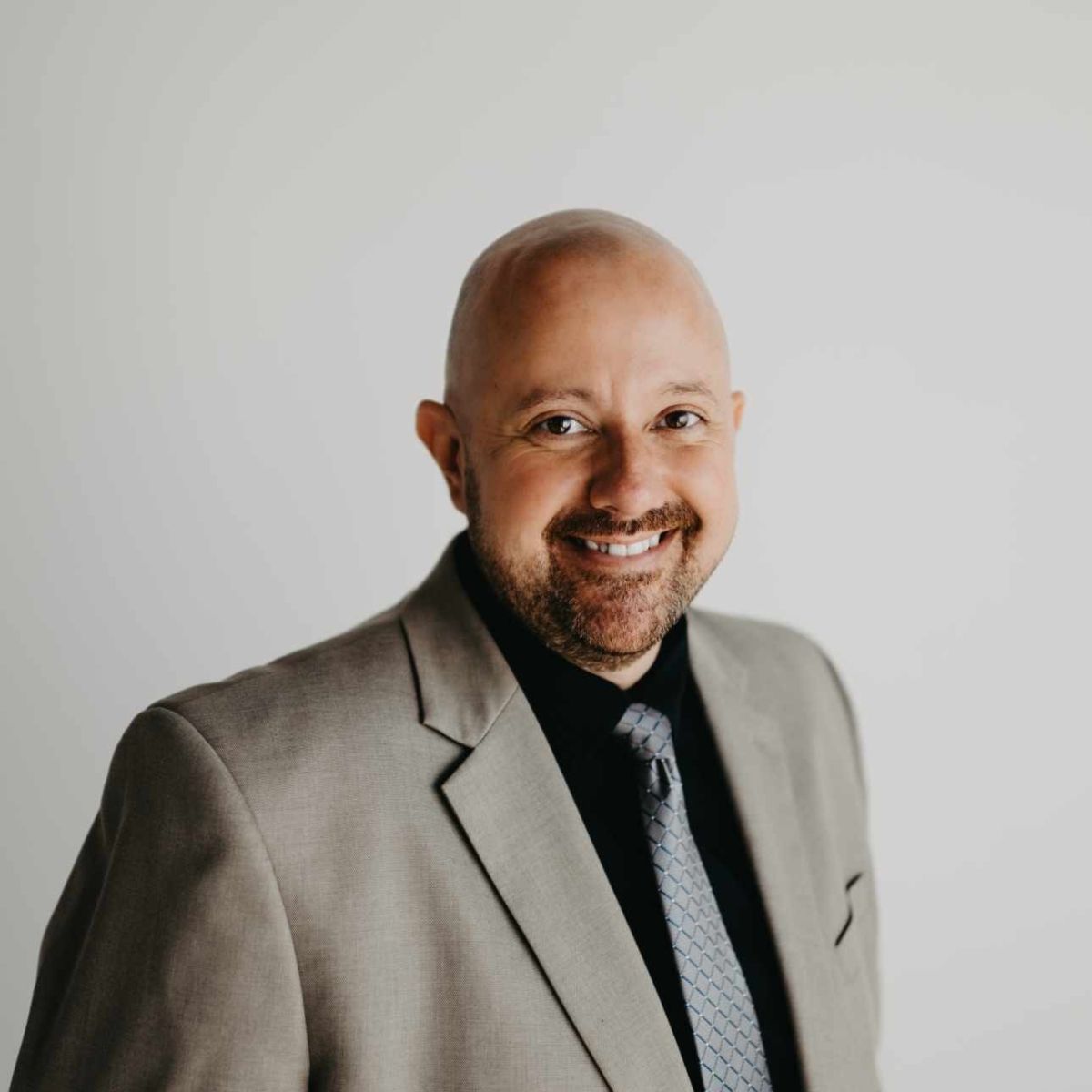 Man with shaved head, smiling in a light gray suit, black shirt, and patterned tie. Against a white background.