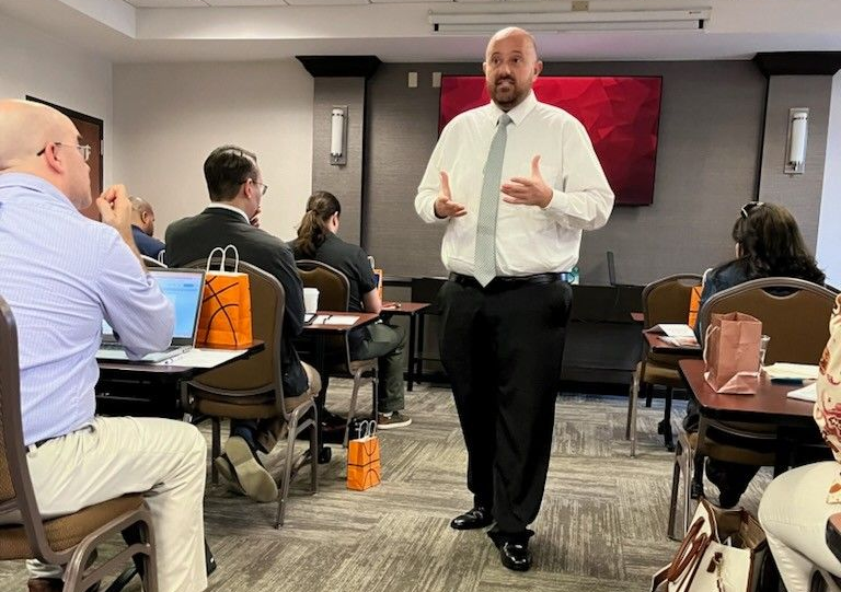 Man speaking in a conference room; gesturing with hands. Audience members seated at tables, some using laptops.