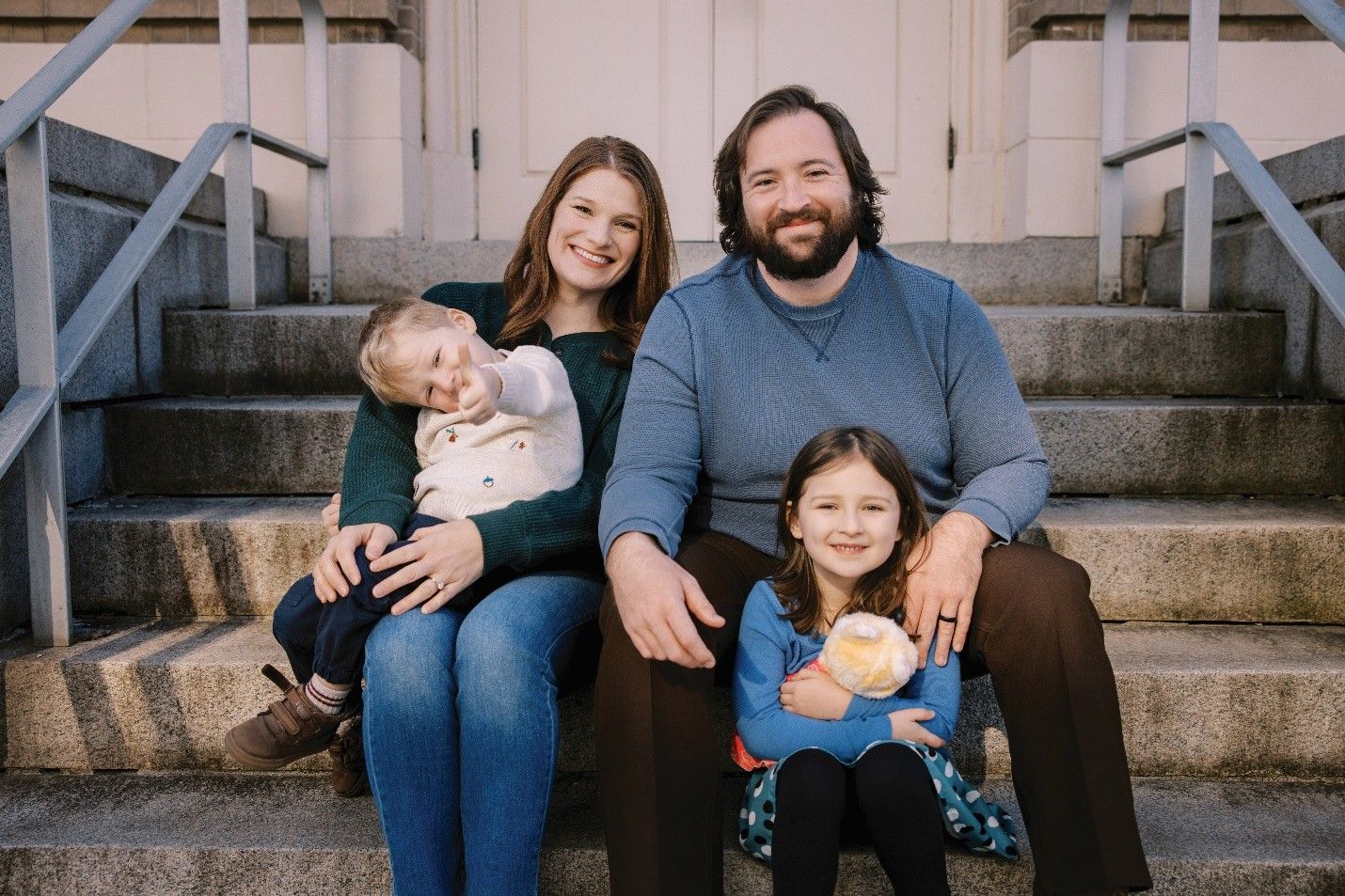 Family of four sitting on concrete steps, smiling. Mother holds young child, girl holds stuffed animal.