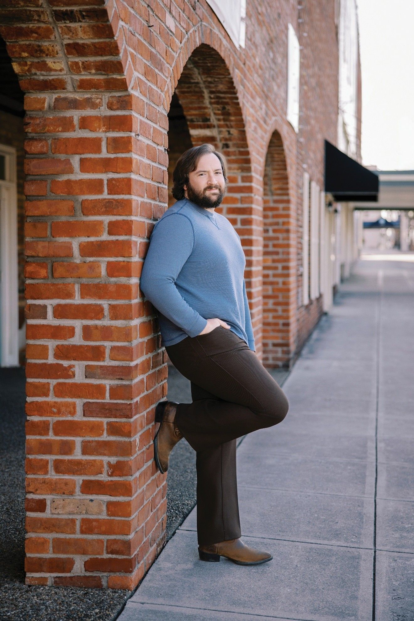 Man in blue sweater and brown pants leans against a brick wall with arched openings.