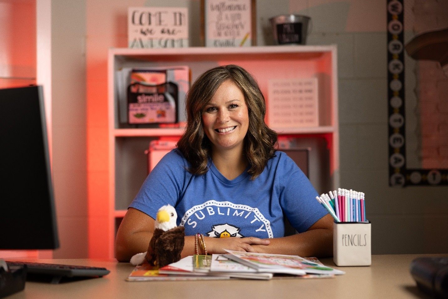 Woman smiling, seated at desk with eagle figurine, pencils, and documents. Blue shirt, bookshelves in background.