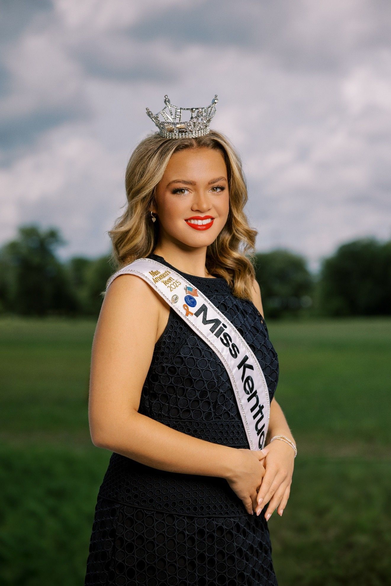 Miss Kentucky, wearing a crown and sash, posing outdoors in front of a field and cloudy sky.