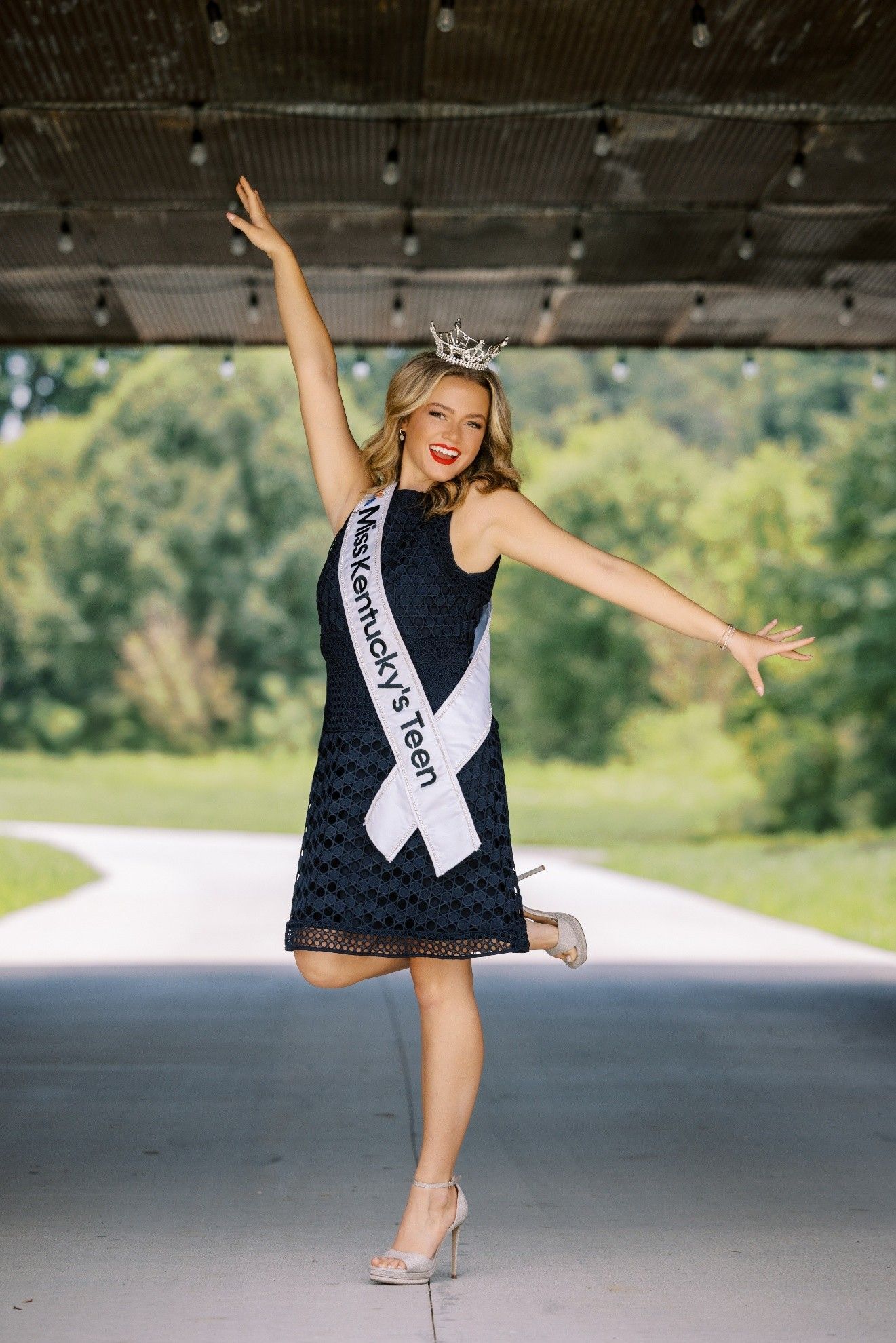 Woman in crown and sash poses with raised arms. Blue dress, heels, outdoor setting.