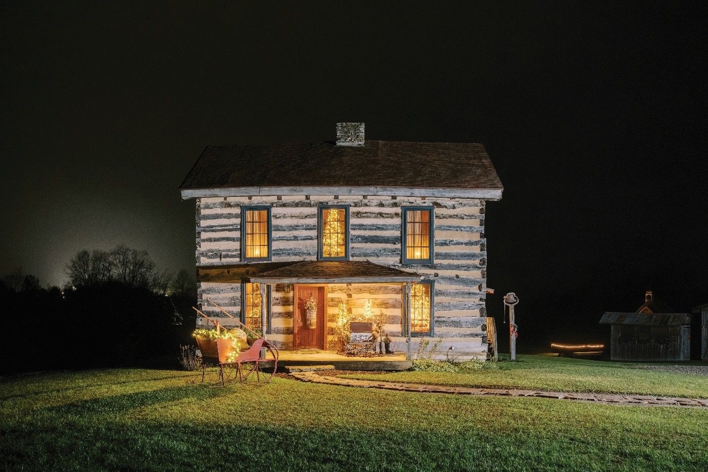 Log cabin at night, illuminated by interior lights, porch glow, long grass, dark sky.