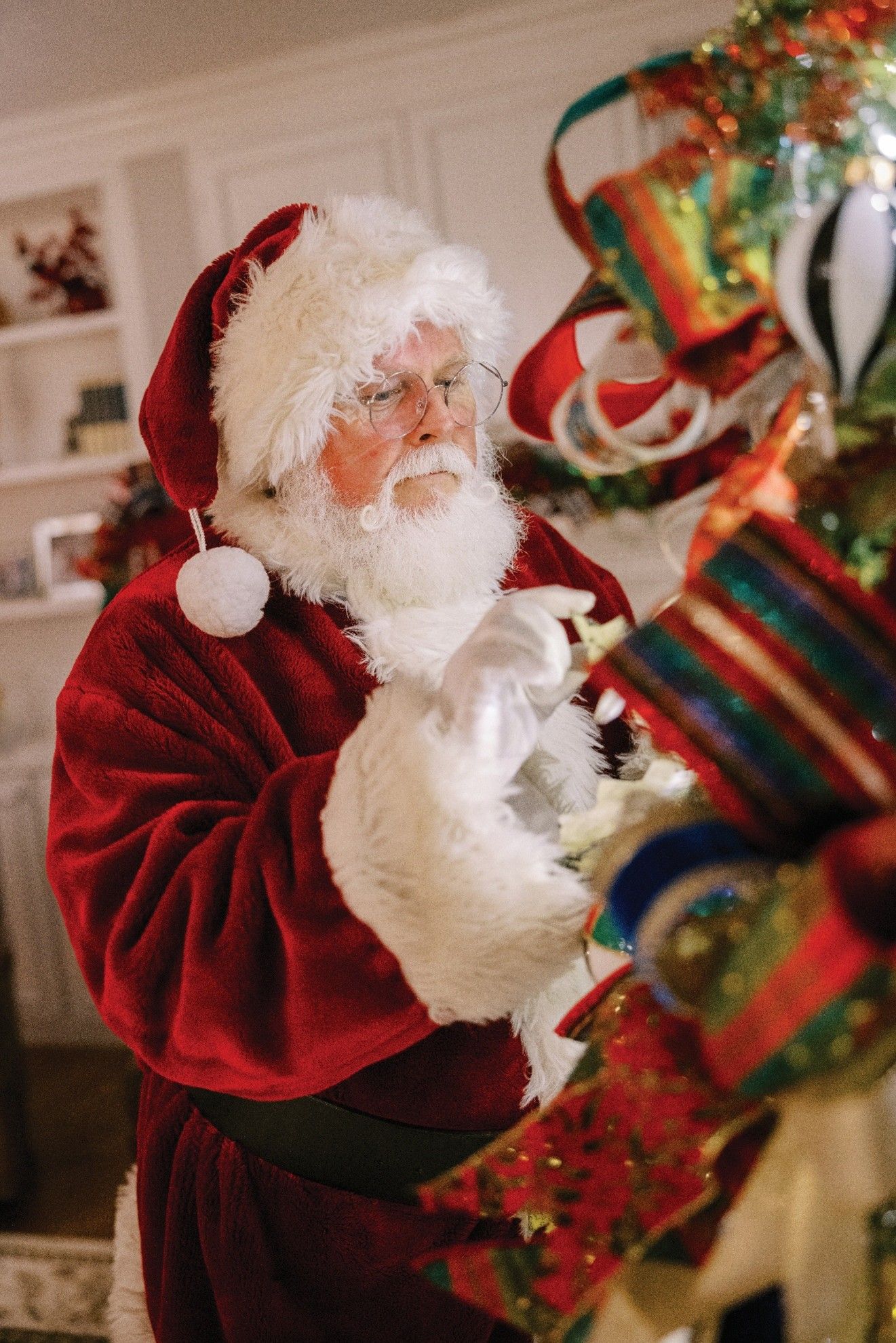 Santa Claus decorating a Christmas tree with red and gold ornaments in a well-lit living room.