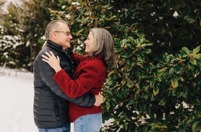 Couple hugging outdoors near evergreen foliage, smiling at each other. Snowy background.