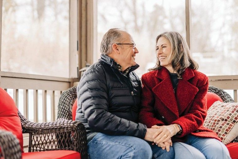 Couple holding hands and smiling on a porch, winter setting. Man in black jacket, woman in red coat.