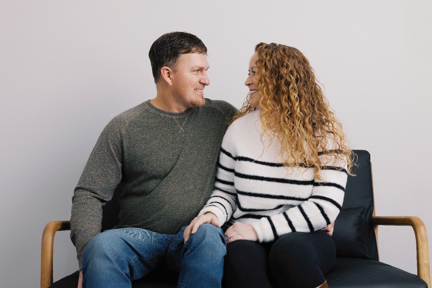 Couple smiling and looking at each other on a modern black couch, neutral background.