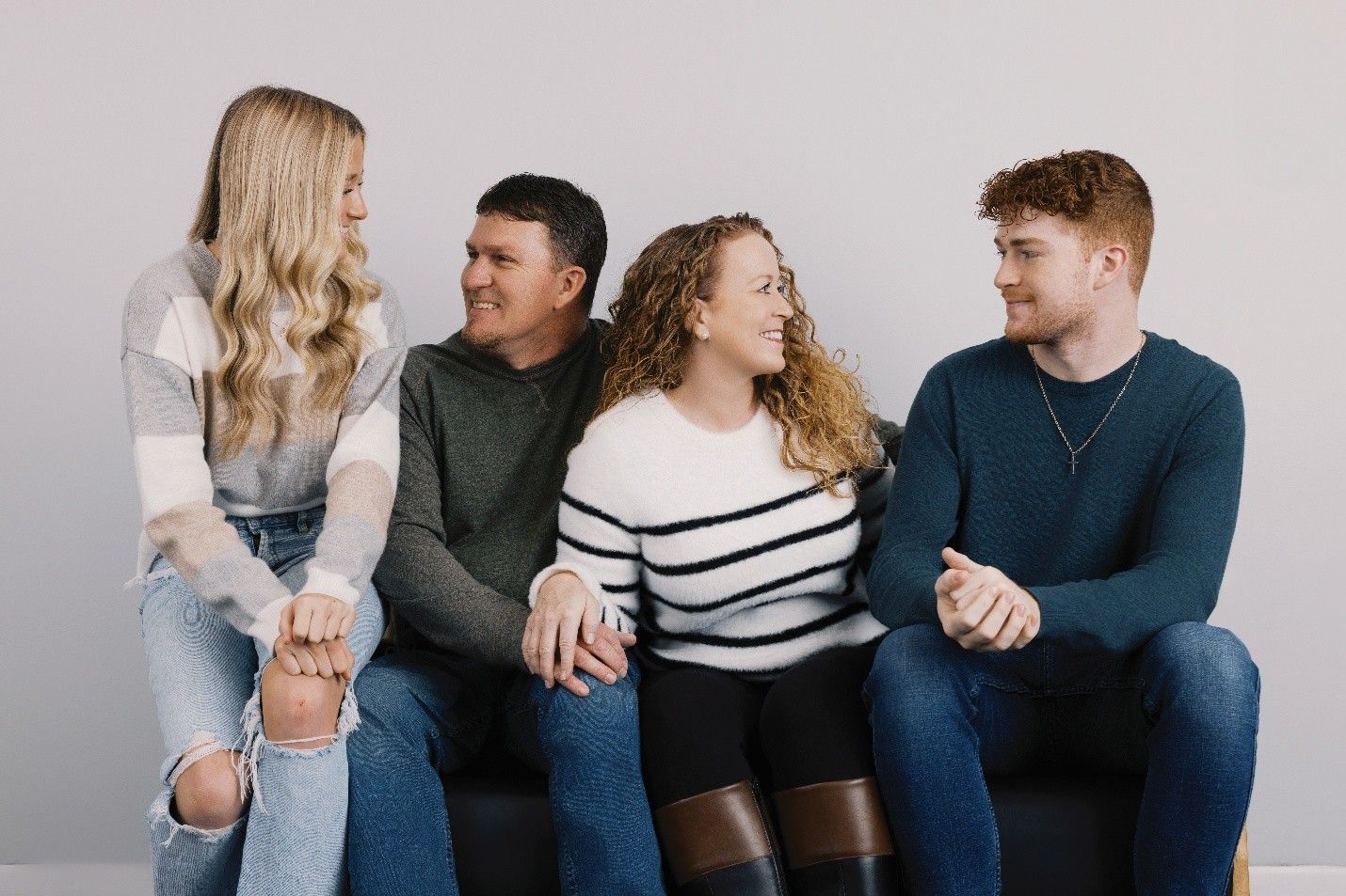 Four people seated together, smiling. Two women, two men, looking at each other. Neutral background.