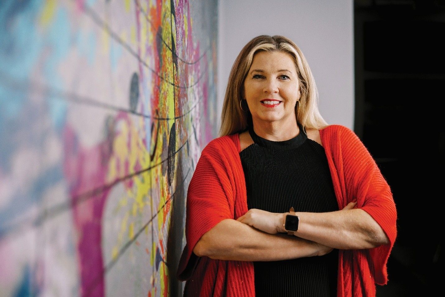 Woman in red cardigan, arms crossed, smiles. Leans on a colorful mural wall, indoor setting.