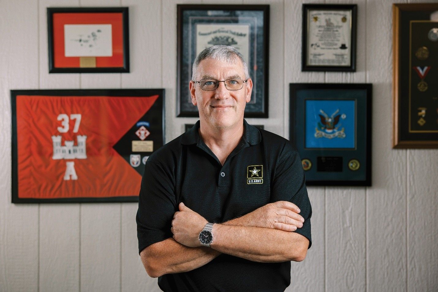 Man in black shirt with arms crossed, in front of framed military memorabilia. White wall.