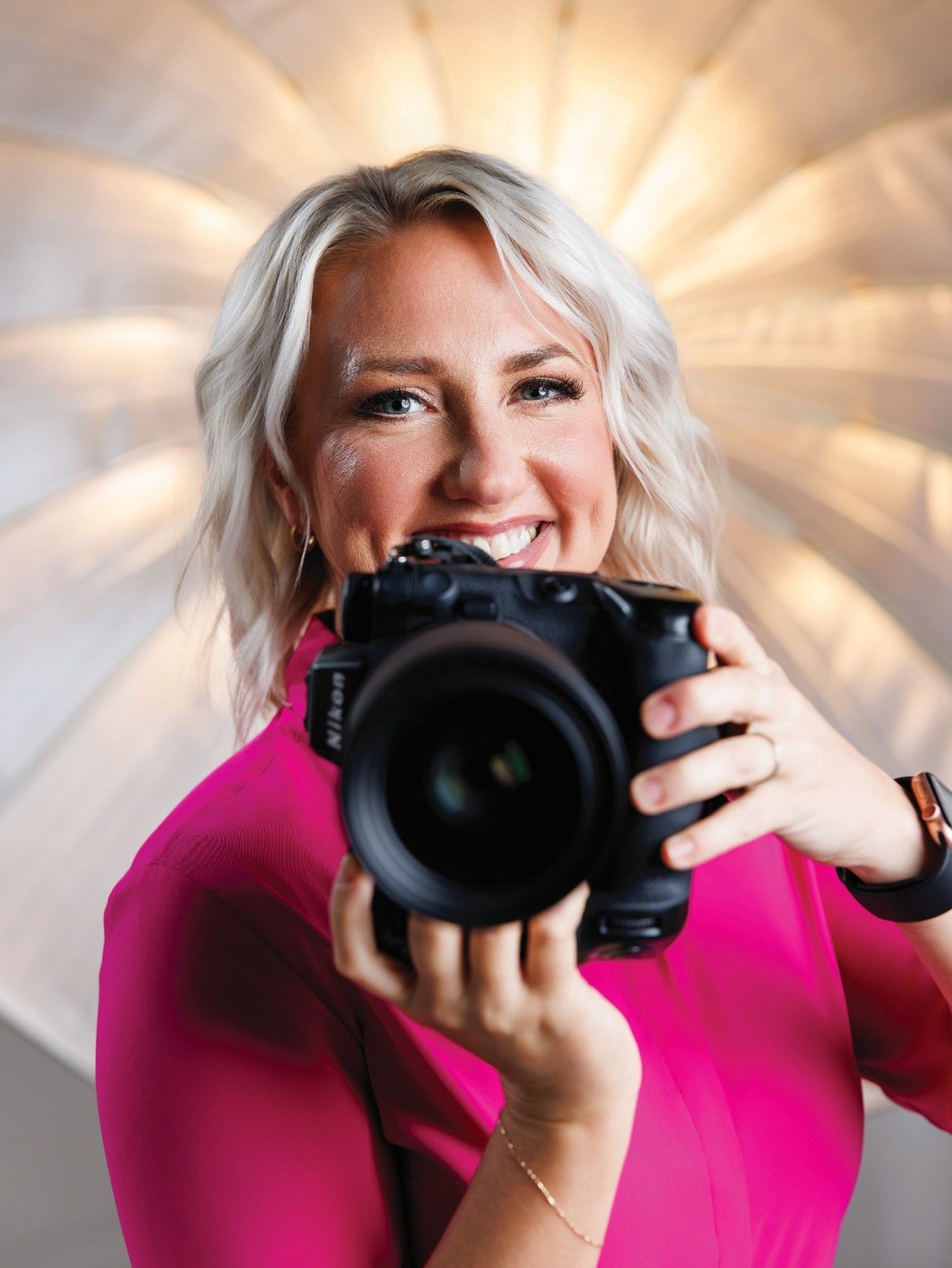 Woman holding a camera, smiling, wearing a pink top, lit by a softbox in a studio.