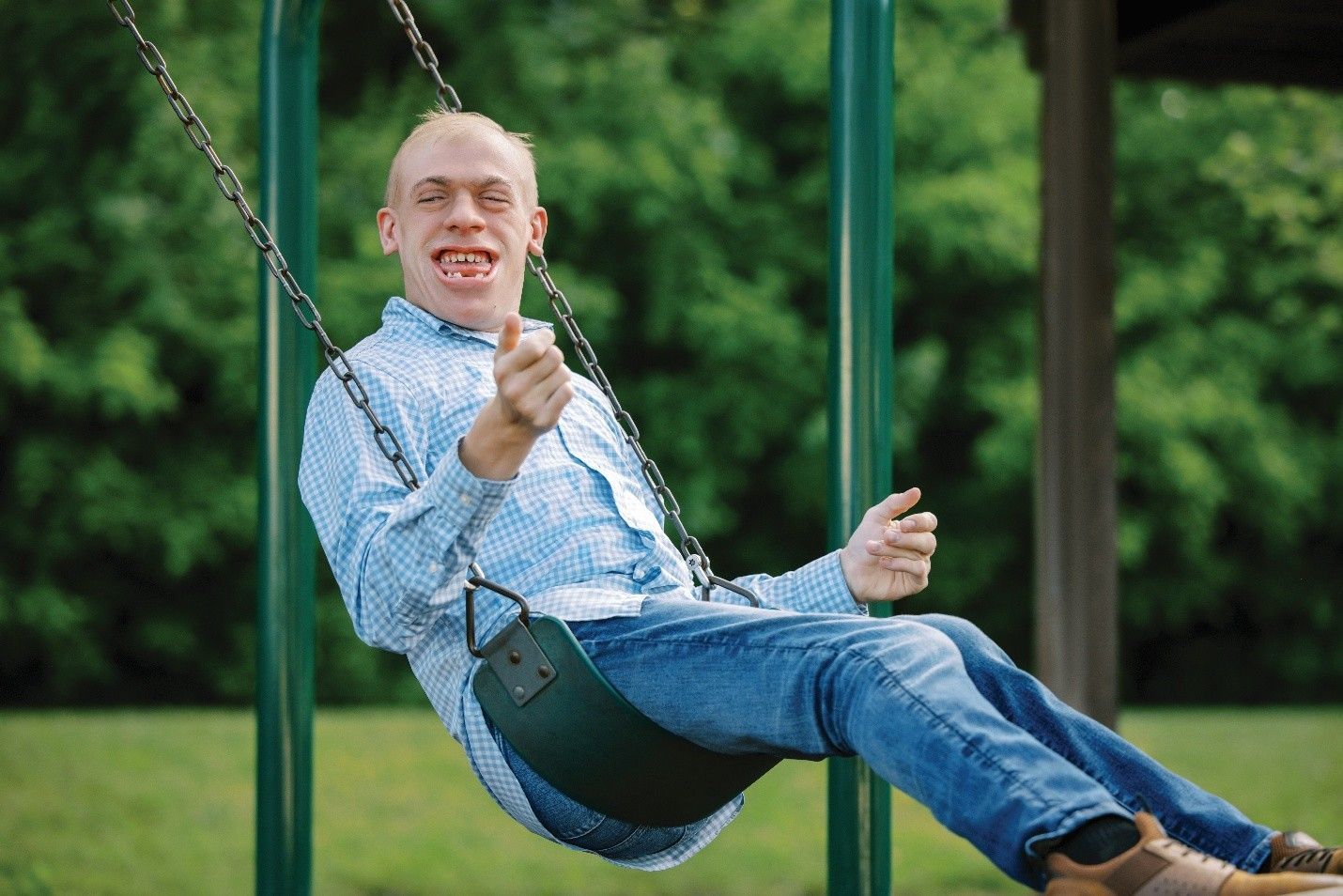 Man on swing smiling, wearing blue jeans and shirt, at park with green swing set.