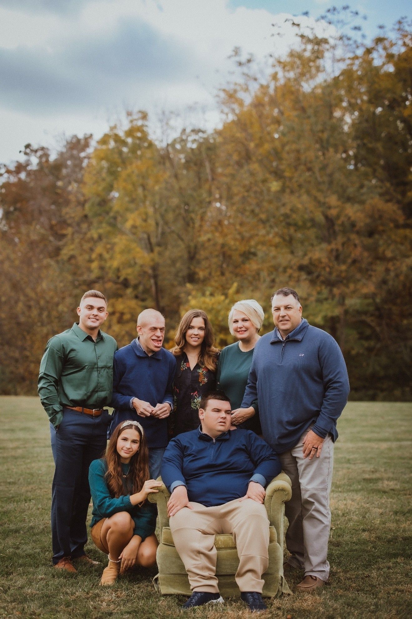 Family portrait outdoors in front of fall foliage. People are smiling. Several wearing blue and green.