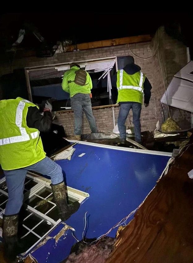 Three people in reflective vests inspect damage to a building, with a broken blue ceiling and debris.