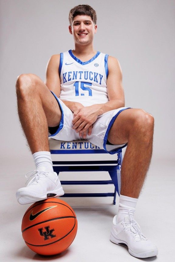 Basketball player in a Kentucky jersey sitting on a blue bench, with a ball at his feet.