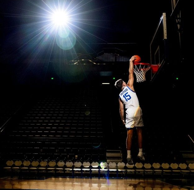 Basketball player in blue uniform dunks ball, arena setting, bright spotlight.