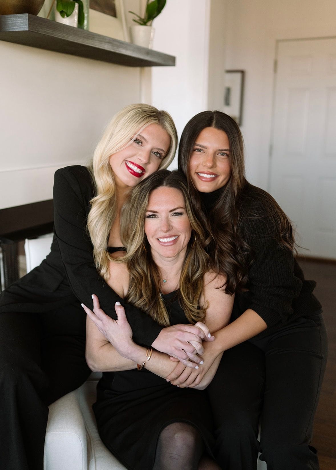 Three smiling individuals in black attire pose together in an indoor setting, with one seated and two standing behind.