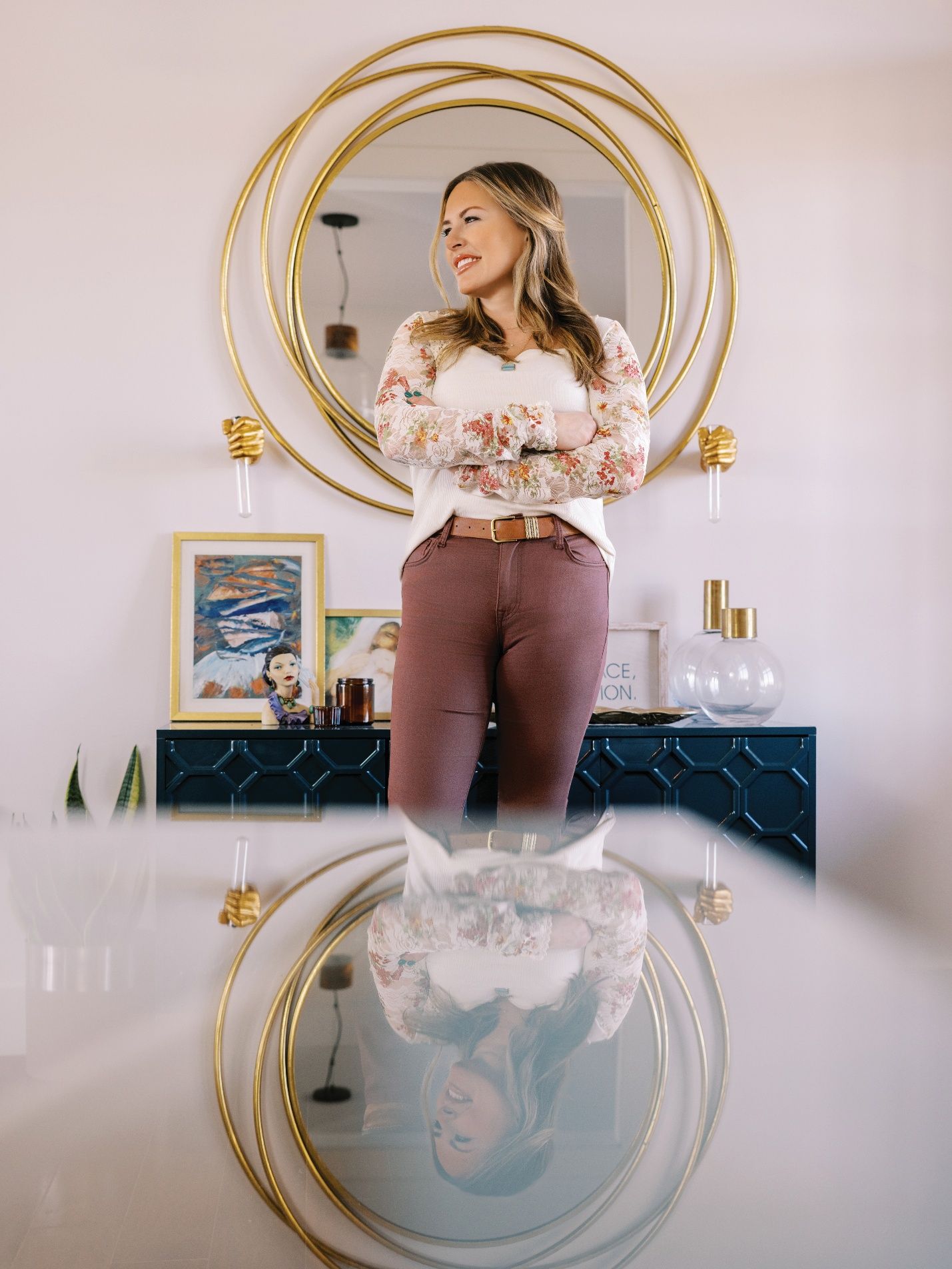 A person stands before a large, round gold mirror, with their reflection visible in the table surface in the foreground.
