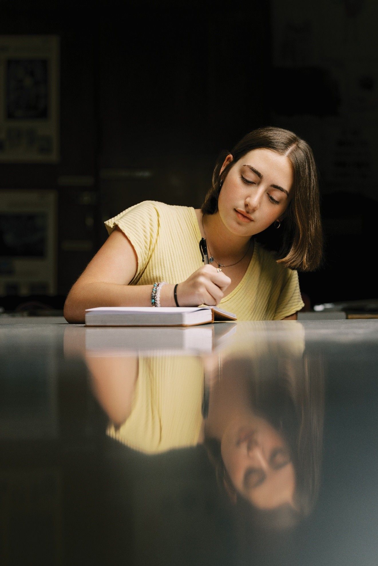 Woman writing in a notebook, reflection on glossy surface. Dark background, yellow top.