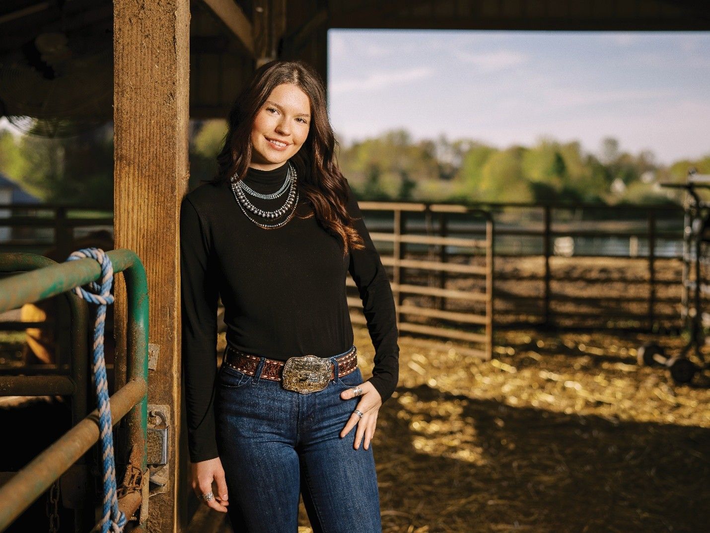 Woman wearing black top, jeans, and belt with buckle, leans against a wooden barn post, smiles.