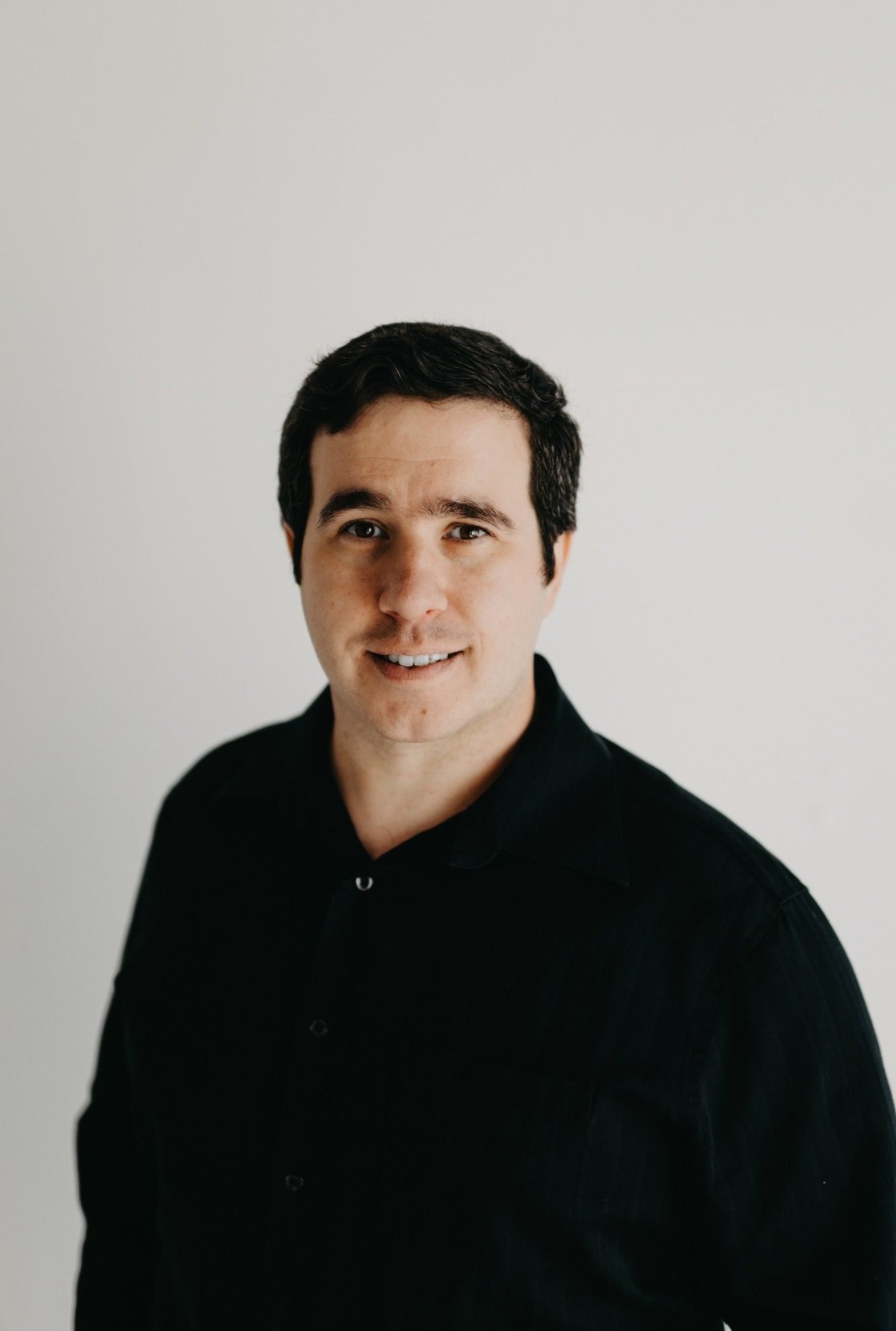 Man with dark hair and a neutral expression wearing a black sweater against a white backdrop.