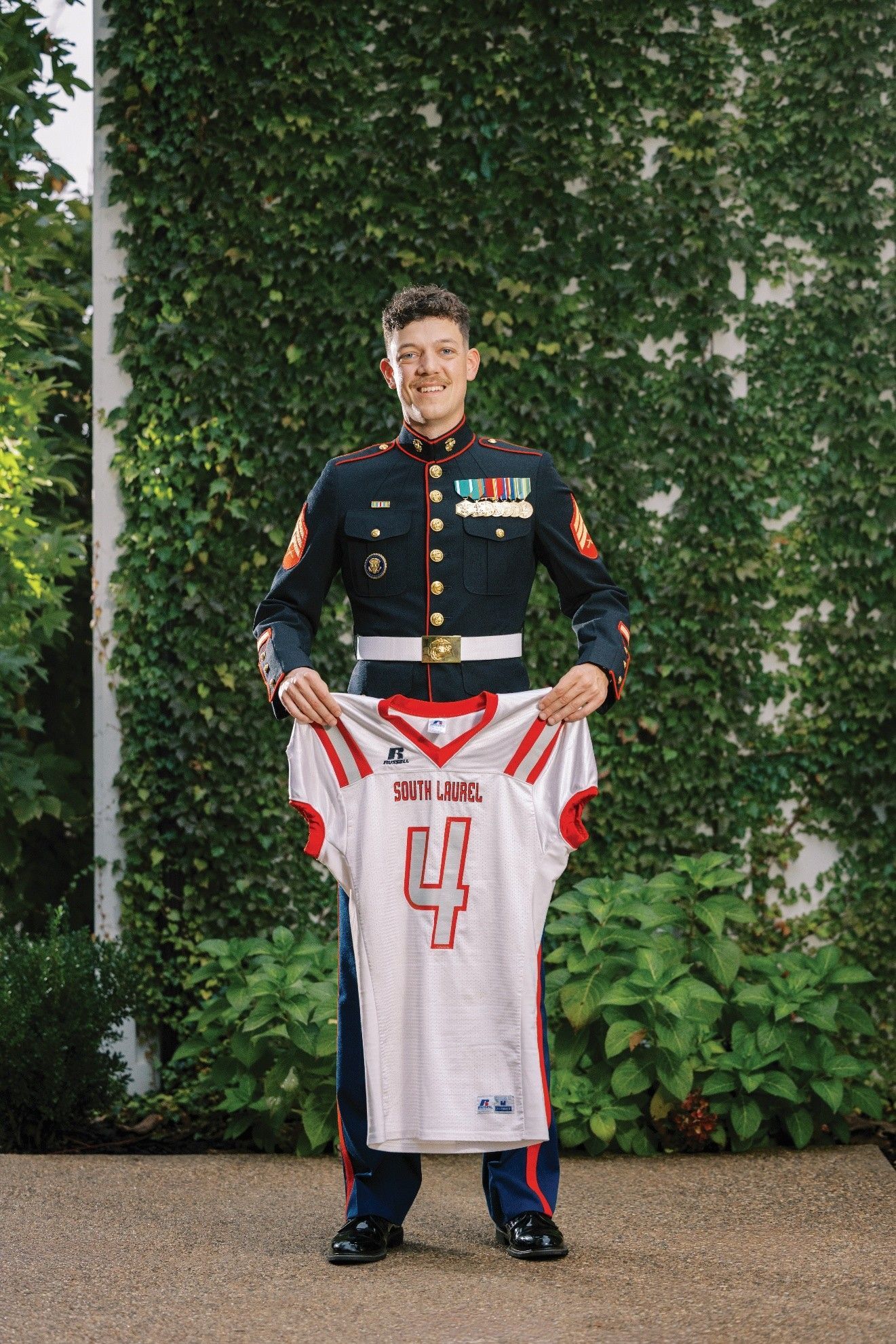 Marine in uniform holding a football jersey with a leafy green backdrop.