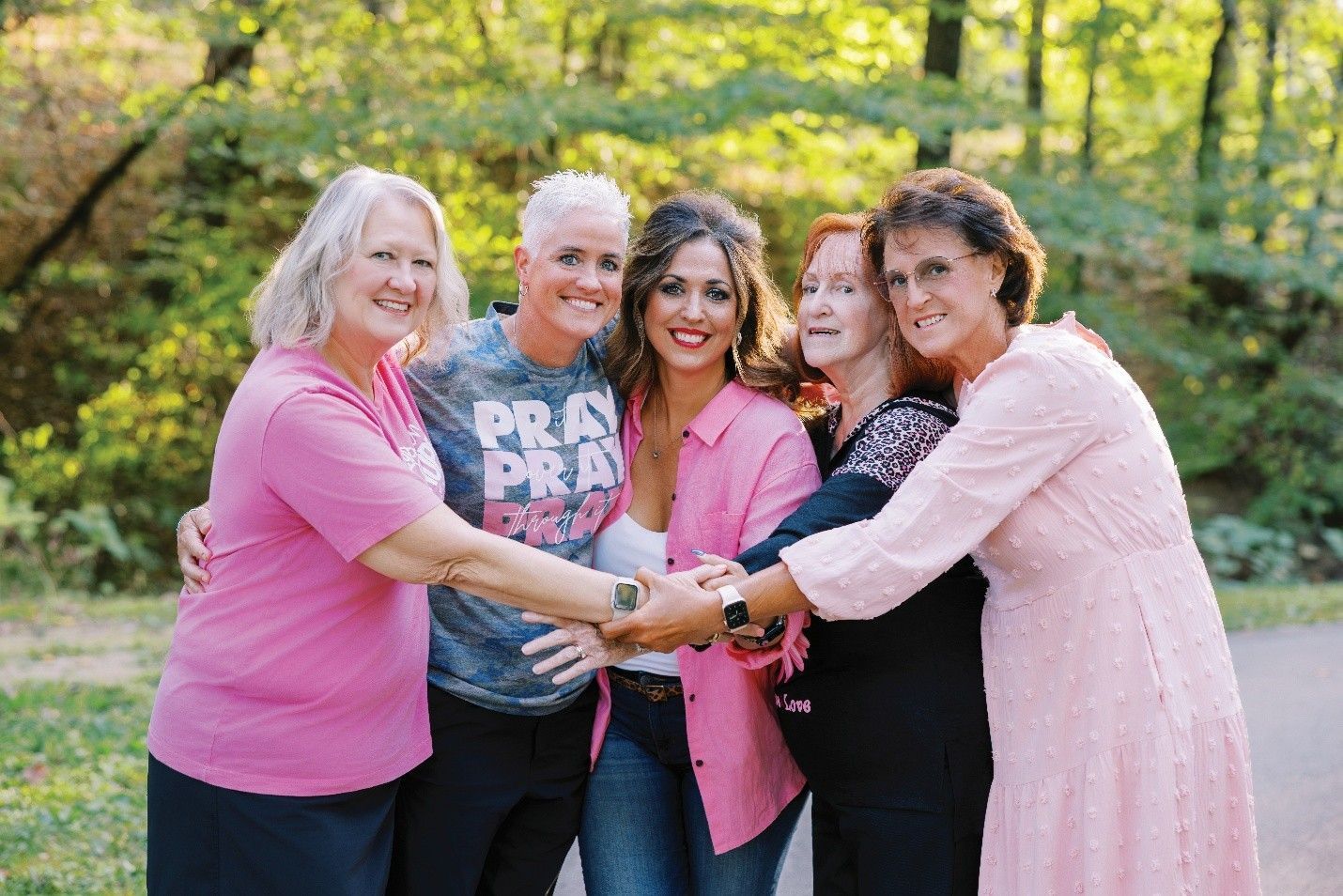 Five people in a park, arms around each other. Pink shirts and a “PRAY” shirt are visible.