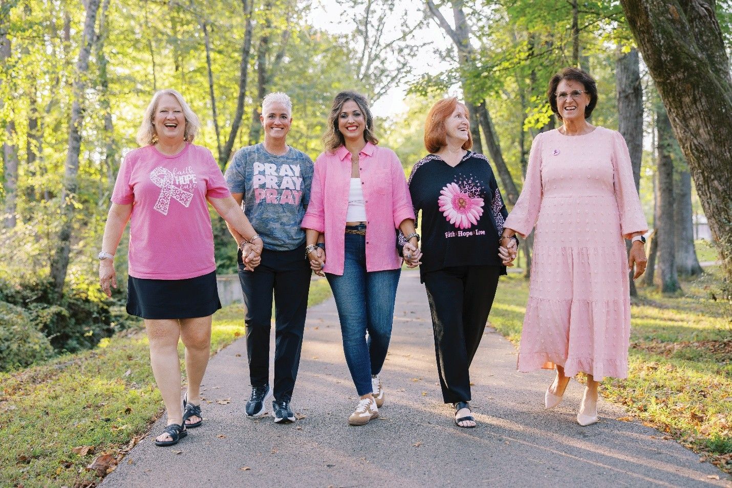 Five women in pink clothing hold hands, walking outdoors.