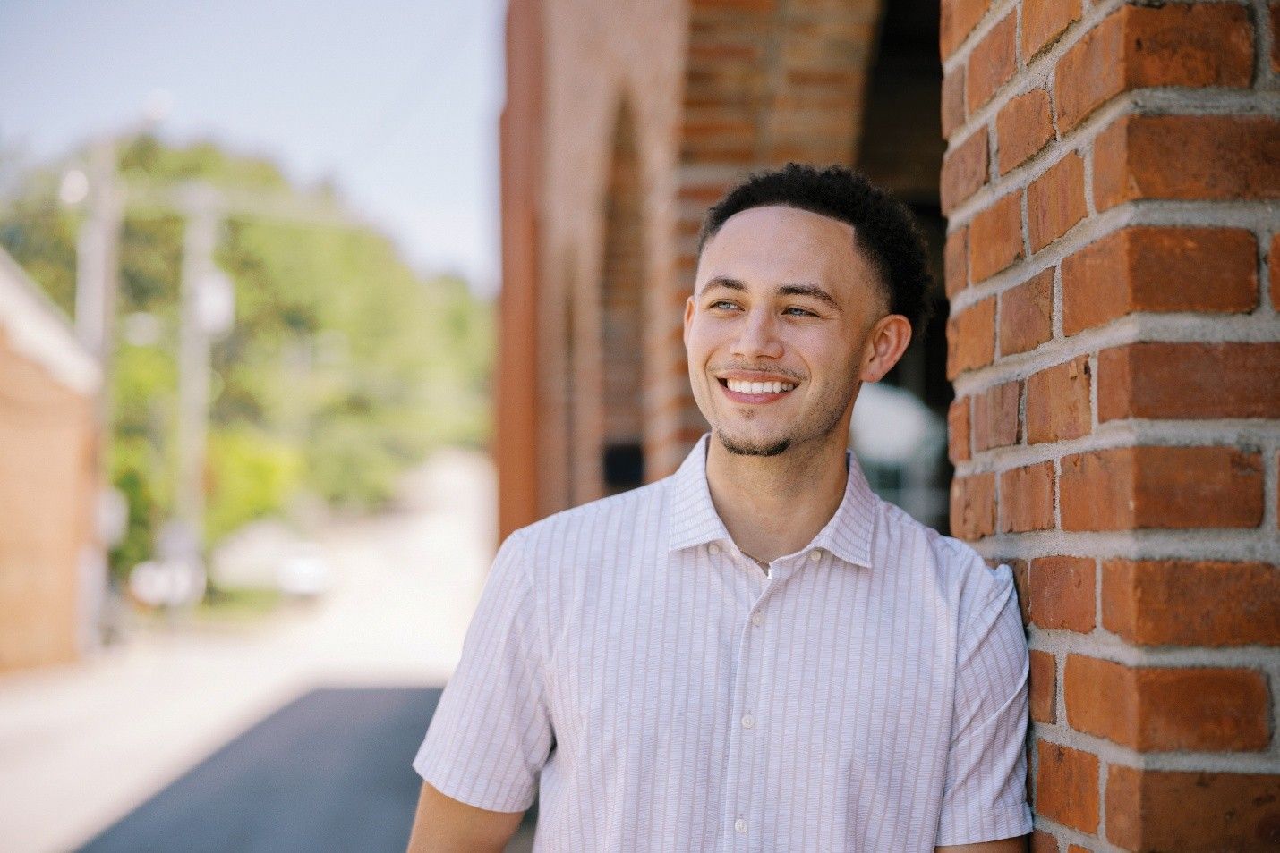 Man in a light pink shirt smiles next to a brick building outdoors.