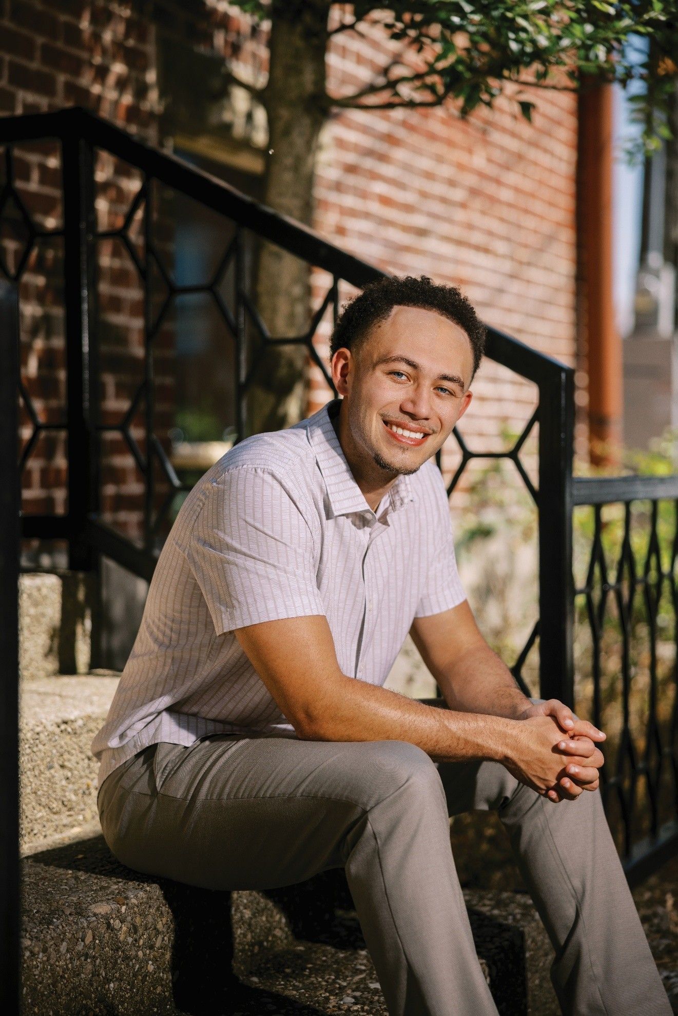 Man sits smiling on outdoor steps, arms resting. He wears a light patterned shirt and gray pants.