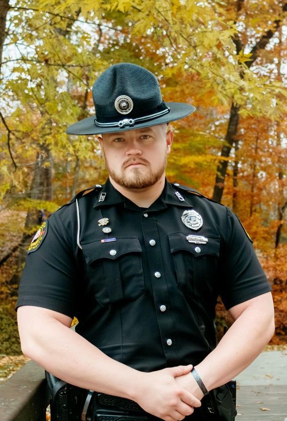 Police officer in black uniform and hat, arms crossed, standing outdoors with autumn foliage.