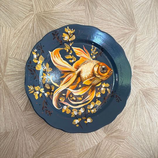 Een grijs handbeschilderd wandbord met een gedetailleerde, goudoranje goudvis, omringd door geschilderde gouden bloemaccenten